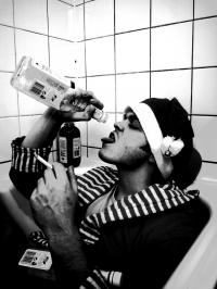 Foto-Abbildung eines Manns, der mit Bademantel und Mütze bekleidet in der Badewanne liegt und Alkoholflaschen im Arm hält. Mit einer Hand führt er sich eine Flasche Alkohol zu, in der anderen Hand hält er eine glimmende Zigarette.
